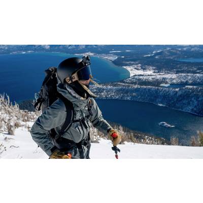 Eine Person mit Skiausrüstung steht auf einem schneebedeckten Hang, mit Blick auf einen weitläufigen See und bewaldetes Land im Hintergrund.