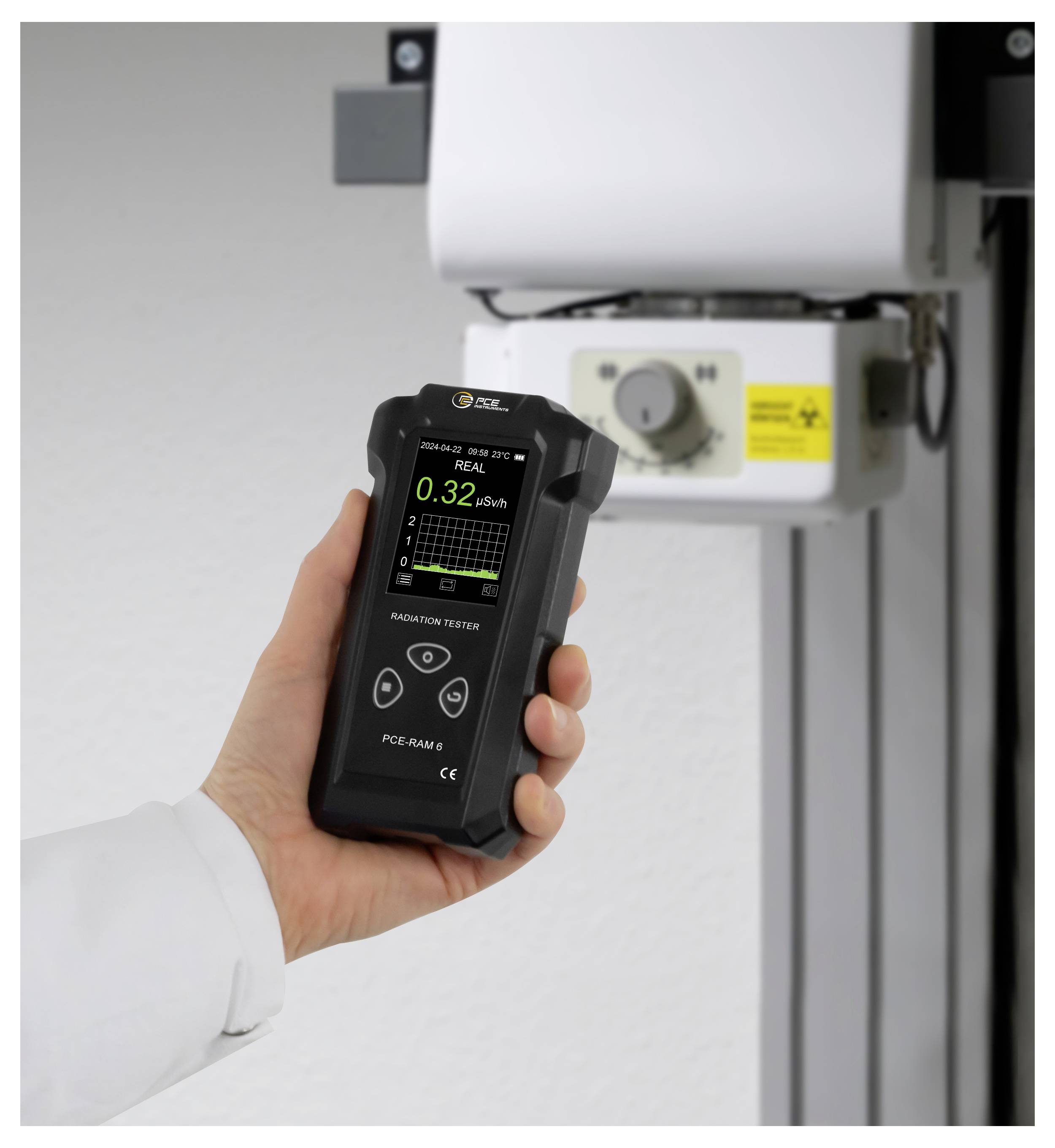 Eine Hand hält ein Strahlenmessgerät in der Nähe von medizinischen Geräten, wobei das Display eine niedrige Messung von 0,32 μSv/h anzeigt.