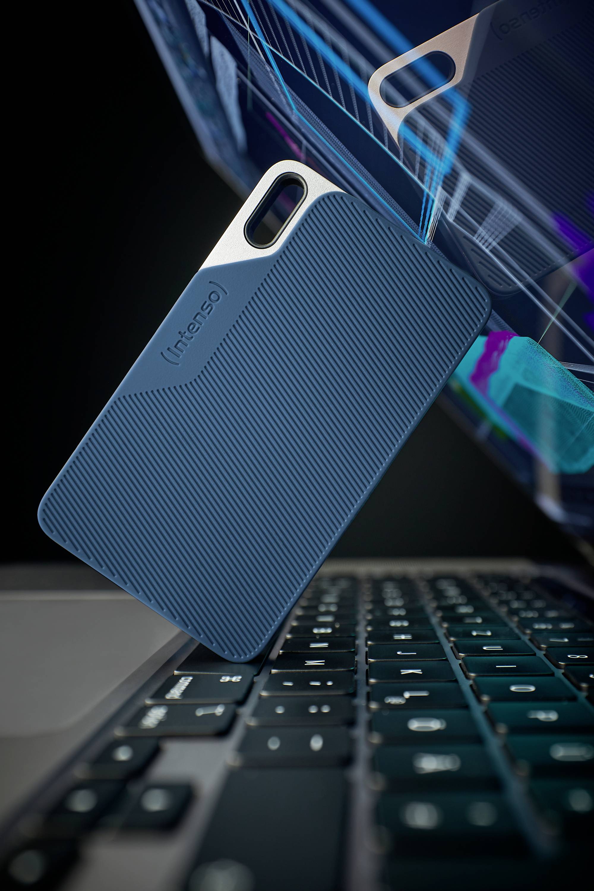 Externe Festplatte lehnt an einem Laptop. Blaues Gehäuse mit strukturiertem Design. Reflektionen und technische Zeichnungen im Hintergrund.