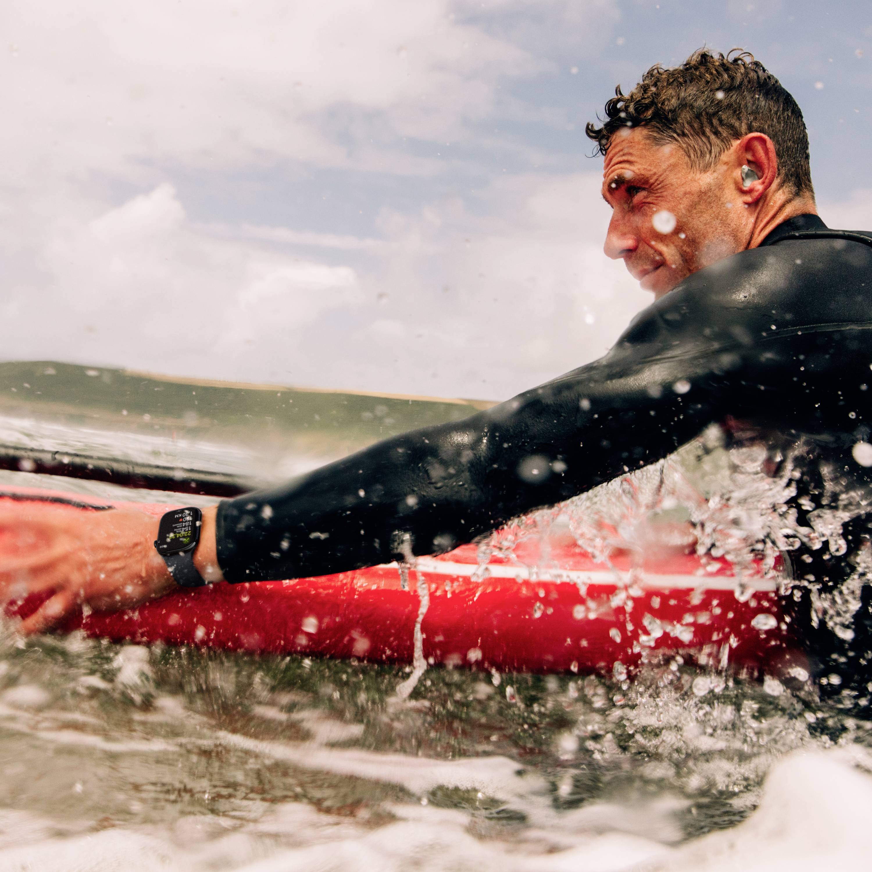 Eine Person surft auf einer Welle mit einem roten Surfbrett, trägt einen schwarzen Neoprenanzug und hat Ohrstöpsel im Ohr.