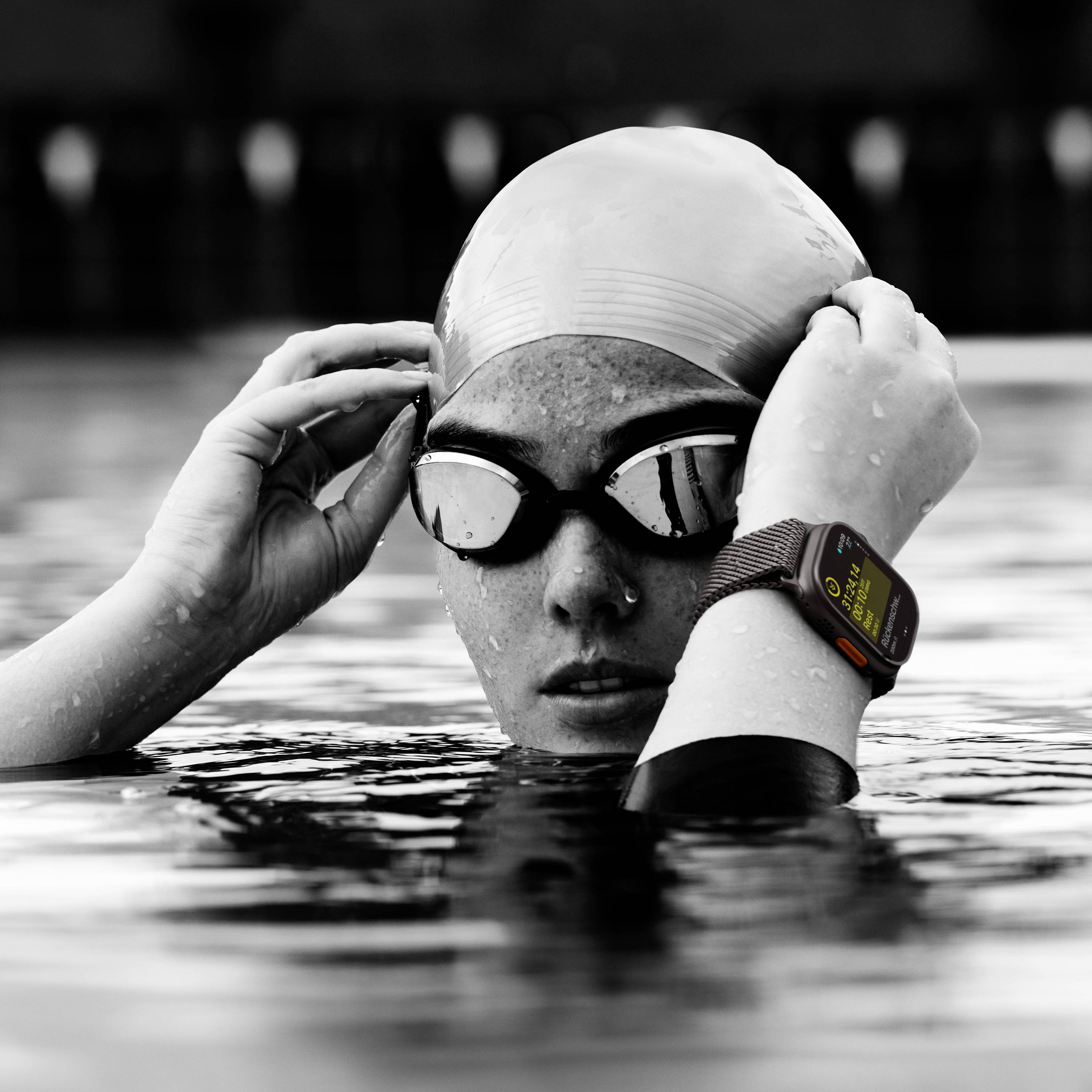 Eine Schwimmerin mit Schwimmbrille und Badekappe, halb im Wasser, trägt eine Smartwatch am Handgelenk, die Fitnessdaten anzeigt.