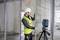 Ein Bauarbeiter mit Schutzhelm und Warnweste justiert einen vertikalen Träger innerhalb einer teilweise errichteten Struktur, wobei in der Nähe ein Lasernivelliergerät auf einem Stativ steht.