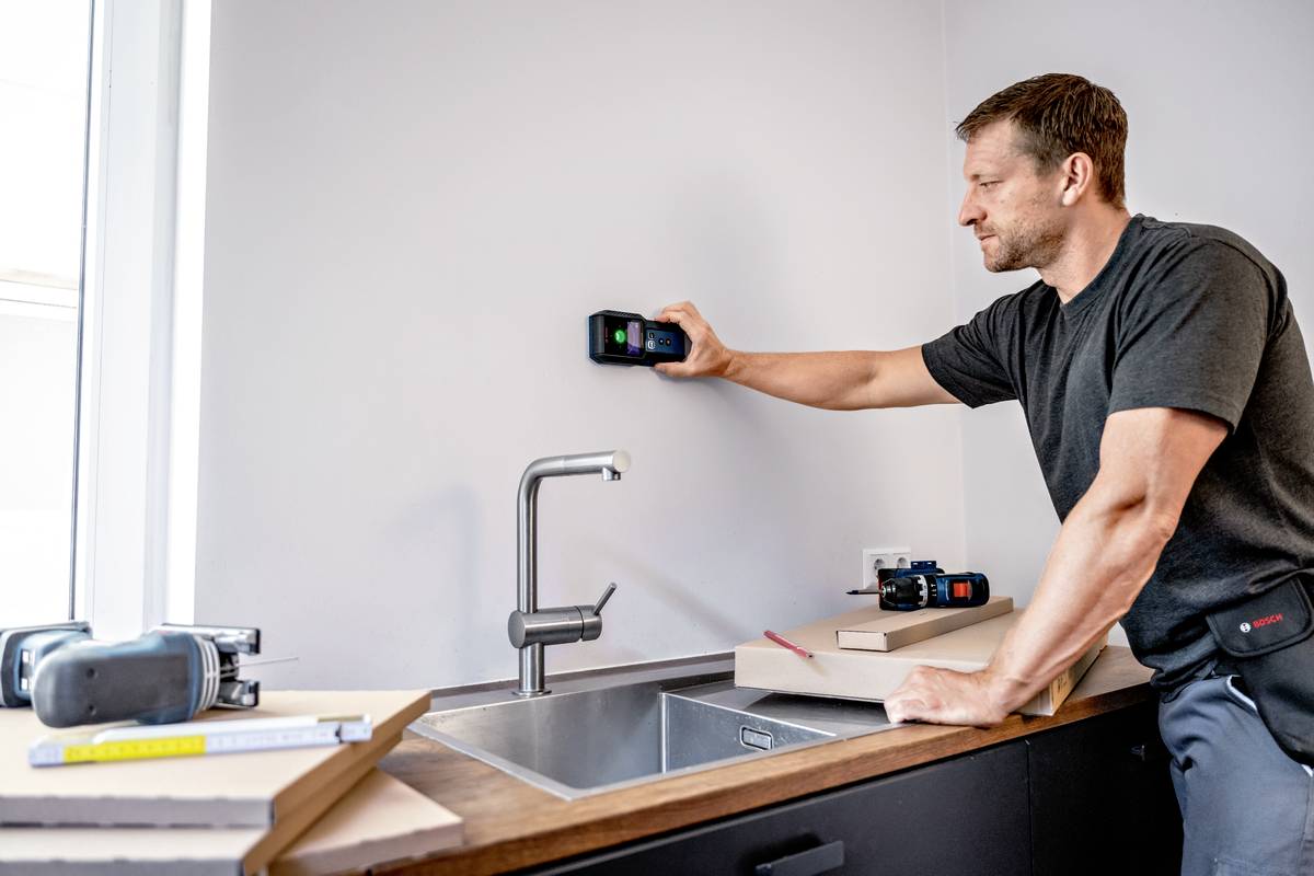 Ein Mann misst mit einem Gerät an der Wand über einer Küchenspüle. Auf der Theke liegen Bücher und Werkzeuge.