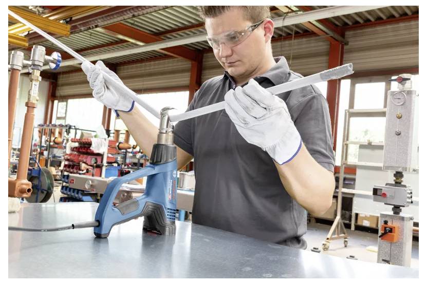 Eine Person trägt Sicherheitshandschuhe und Schutzbrille und verwendet einen blauen Heißluftföhn, um in einer Werkstatt eine Metallstange zu biegen. Verschiedene Werkzeuge und Ausrüstungen sind zu sehen.