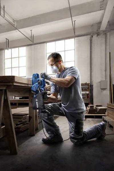 Eine Person mit Sicherheitsausrüstung bedient eine blaue elektrische Säge auf Holzplanken in einer Werkstatt mit hohen Fenstern, wobei eine konzentrierte Umgebung zur Geltung kommt.