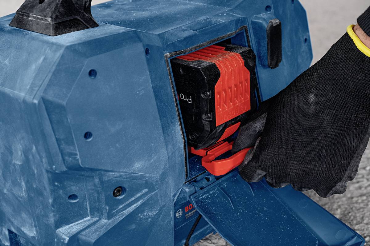 Eine Person mit schwarzem Handschuh wechselt die rote Batterie eines blauen Elektrowerkzeugs mit Staubspuren.