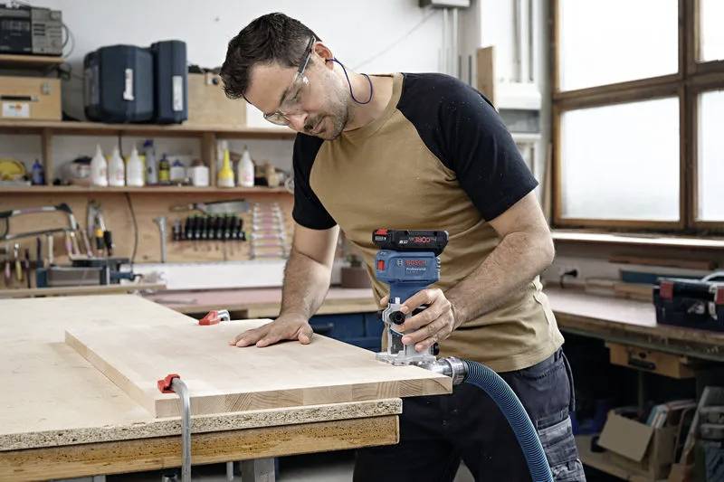 Ein Mann mit Sicherheitsbrille benutzt einen Handrouter auf einem Holzbrett in einer Werkstatt und zeigt dabei eine Holzbearbeitungsaktivität und Werkzeugnutzung.