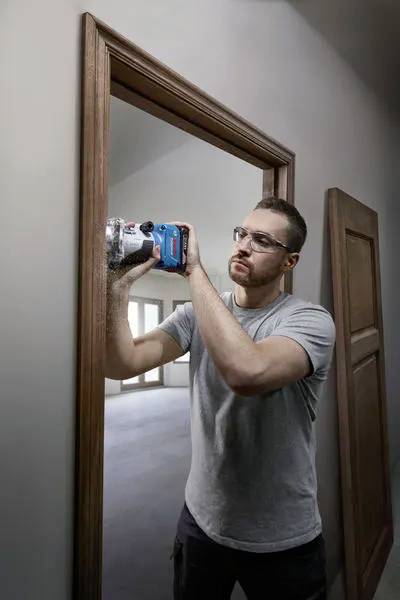 Ein Mann mit Schutzbrille benutzt ein Elektrowerkzeug, um an einem Türrahmen aus Holz in einem Raum zu arbeiten.