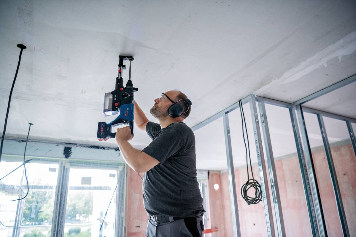 Ein Mann auf einer Baustelle verwendet ein spezielles Werkzeug, um Löcher in eine Decke zu bohren. Er trägt Gehörschutz.