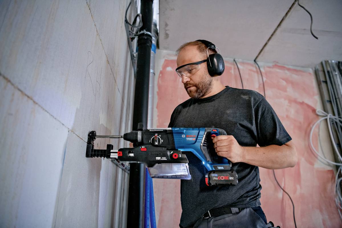 Ein Handwerker mit Schutzbrille und Gehörschutz verwendet eine Bohrmaschine, um ein Loch in eine Wand zu bohren. Hintergrund: Baustelle.