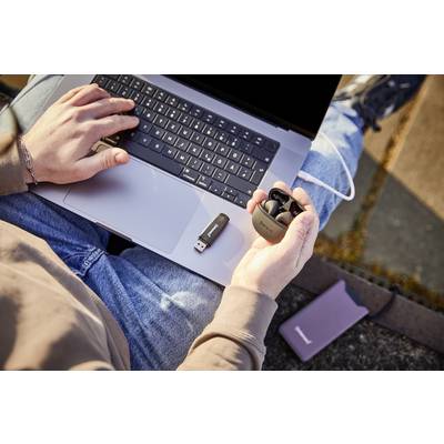 Person sitzt im Freien mit einem Laptop auf dem Schoß, hält kabellose Ohrhörer in der Hand und hat einen USB-Stick daneben liegen.