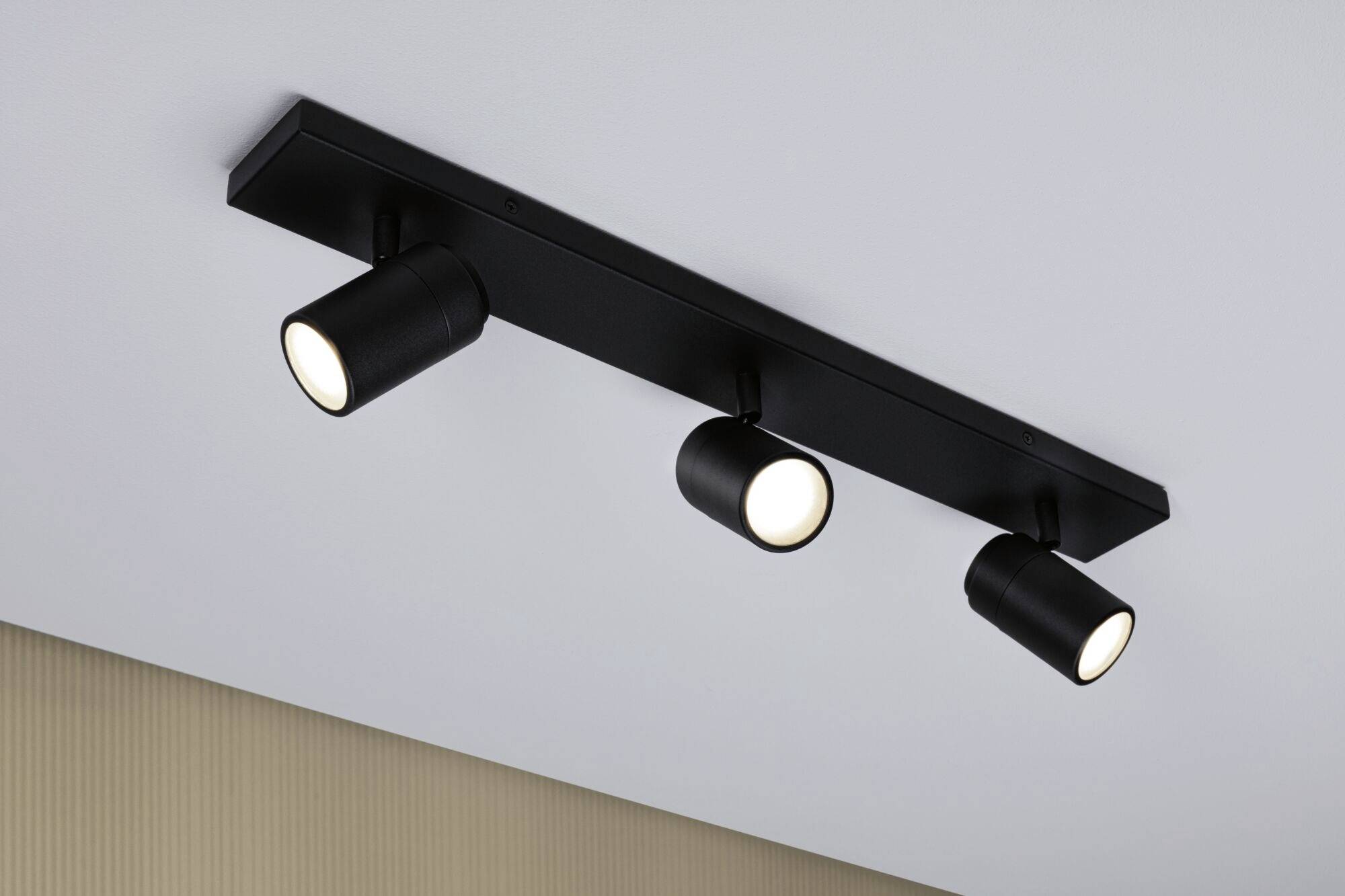 Eine schwarze dreifach-Spotlampe ist an einer weißen Decke montiert. Alle drei Spots sind eingeschaltet und leuchten nach unten.