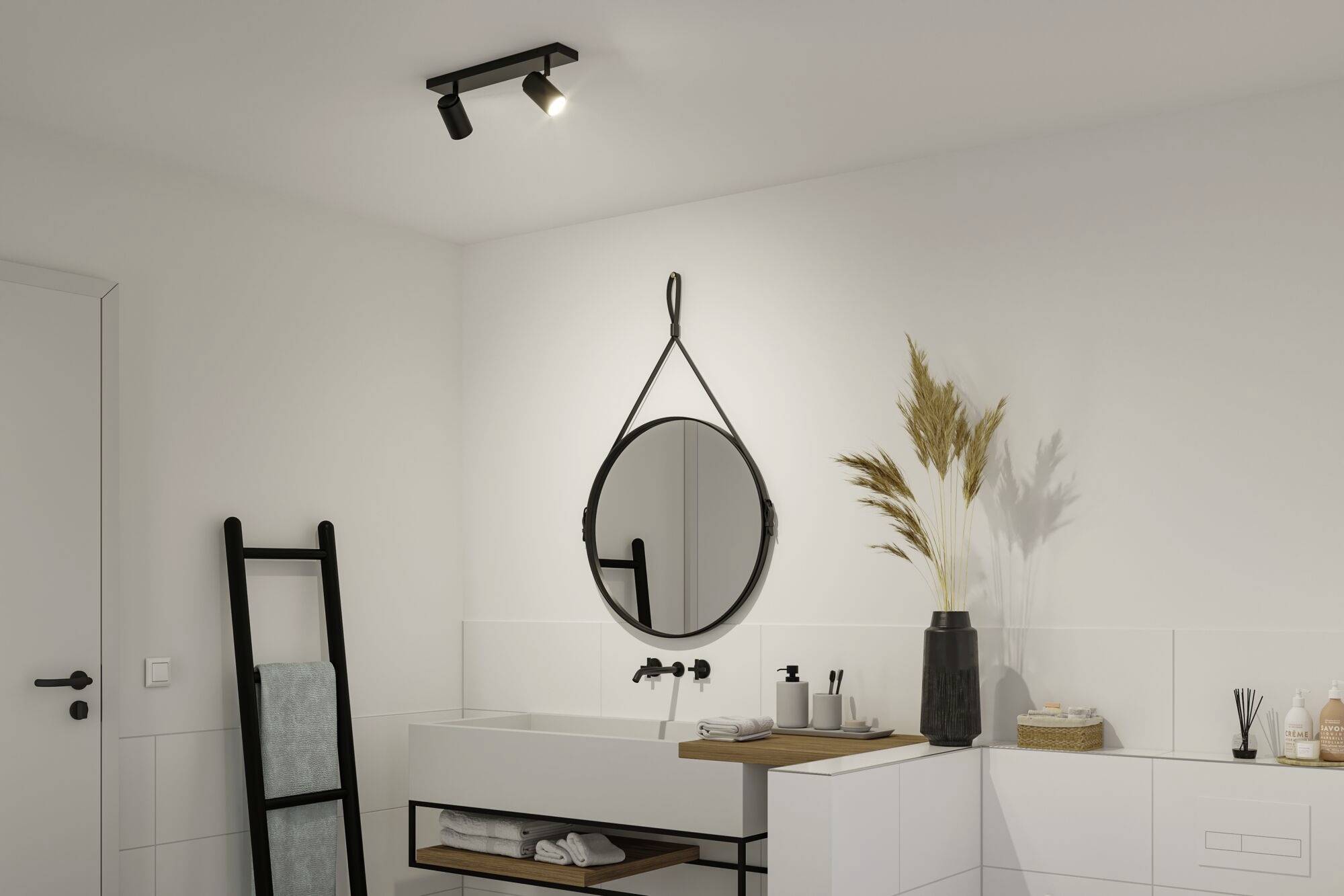 Ein minimalistisch gestaltetes Badezimmer mit rundem Spiegel, Waschbecken, schwarzem Handtuchhalter und dekorativen Gräsern in einer Vase.