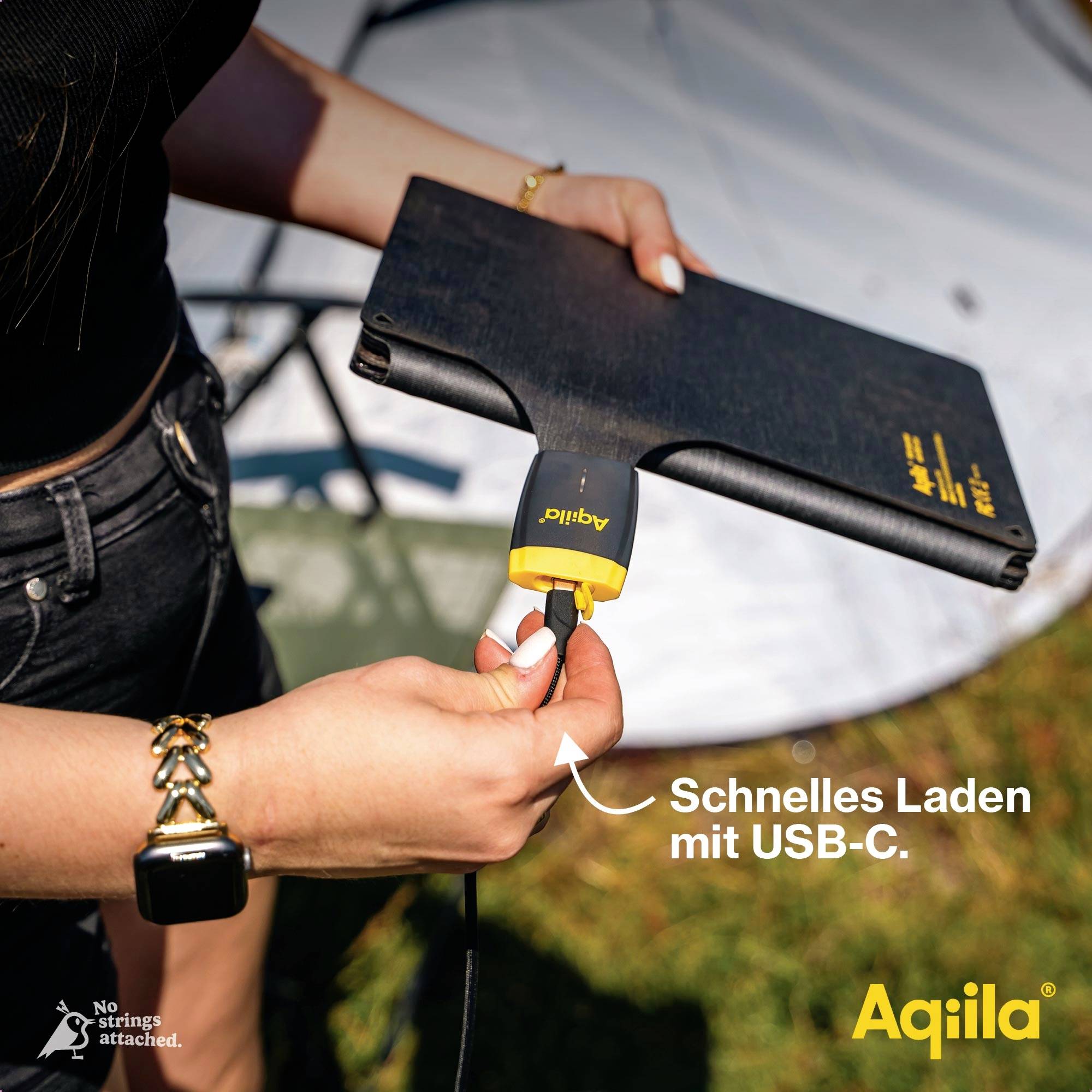Eine Person steckt ein USB-C-Kabel in ein solares Ladegerät mit der Aufschrift 'Schnelles Laden mit USB-C'. Im Hintergrund ist ein Zelt.
