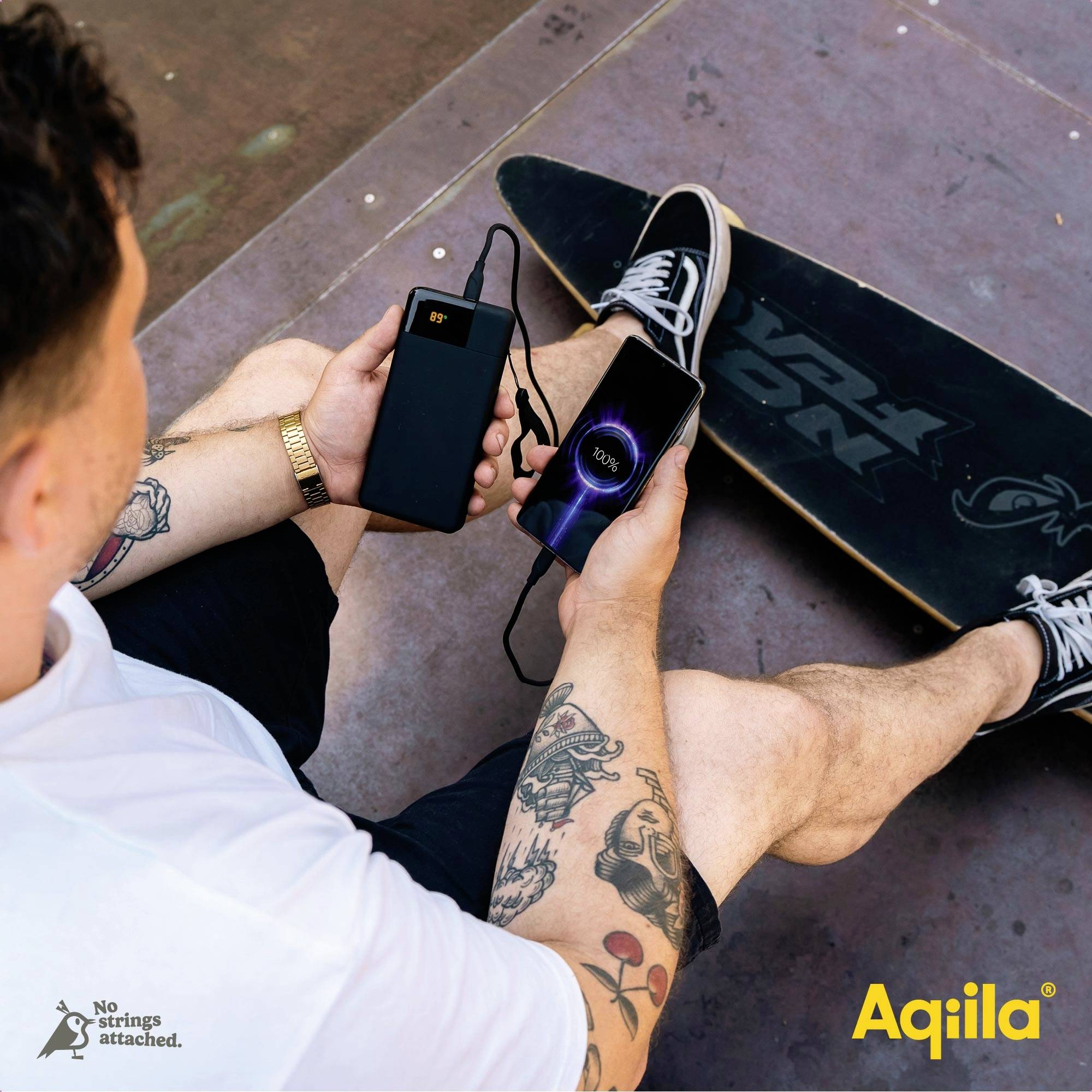 Ein Mann sitzt auf einem Skateboard und lädt sein Handy mit einer Powerbank auf. Text: 'No Strings Attached'. Marke: Aquila.