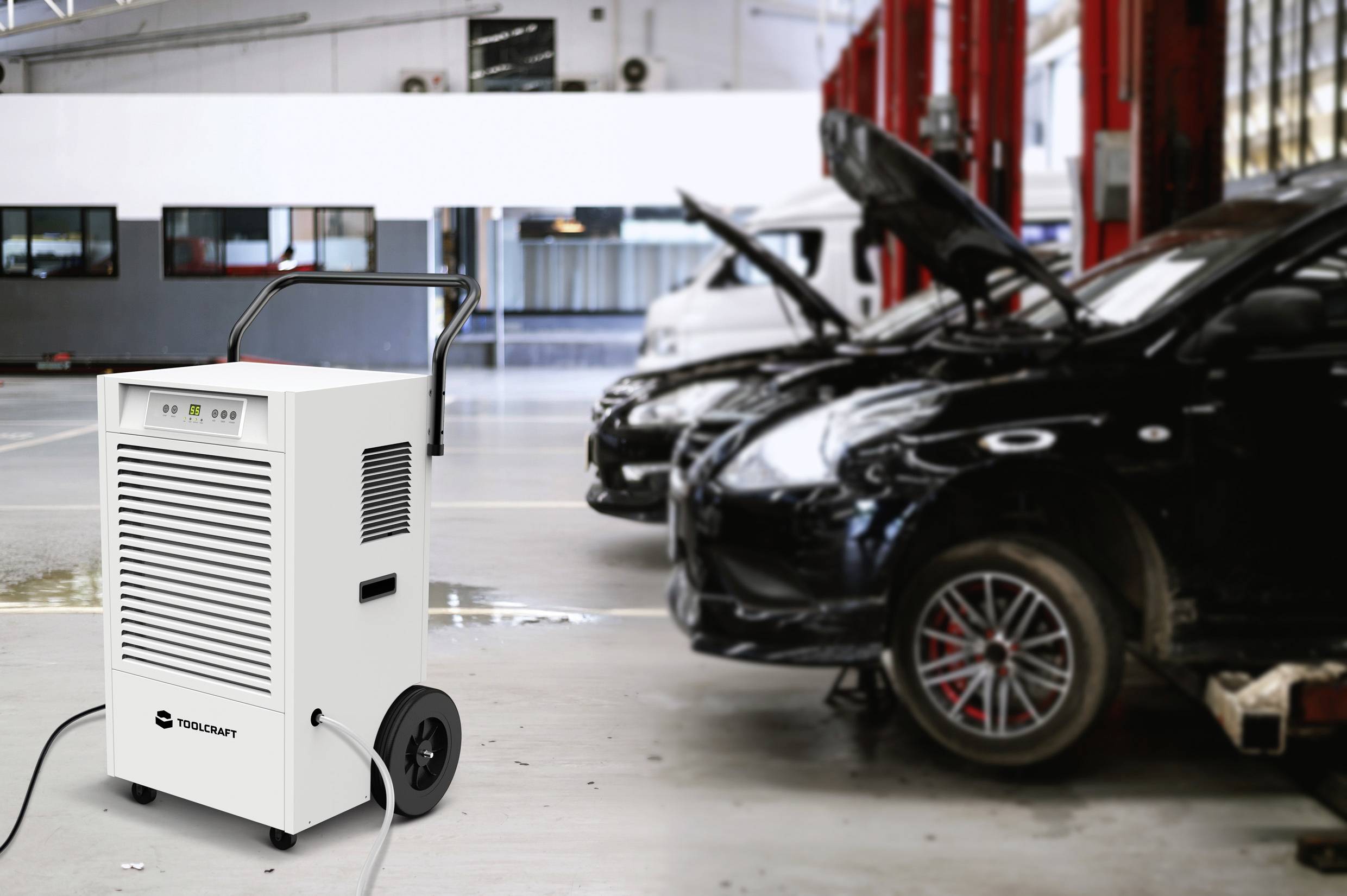 Luftentfeuchter auf Rollen in Werkstatt vor geöffneten Autokappen, bereit zur Feuchtigkeitsregulierung, im Hintergrund Hebebühnen.