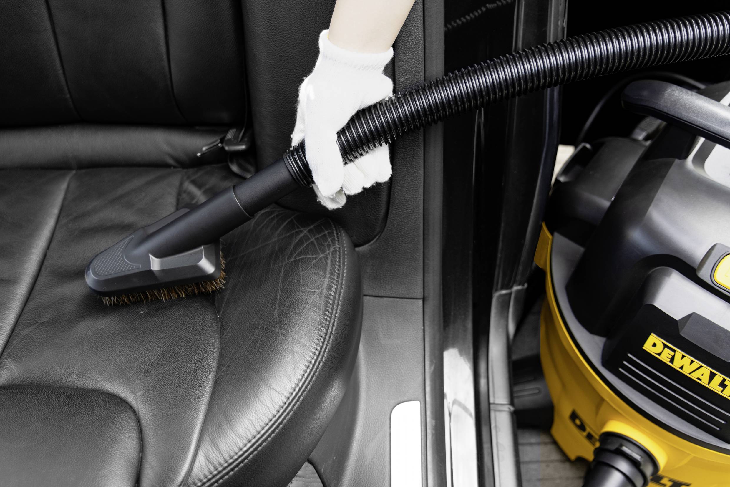 Eine Person reinigt mit einem Staubsaugeraufsatz den Ledersitz eines Autos. Ein gelber Staubsauger steht daneben.
