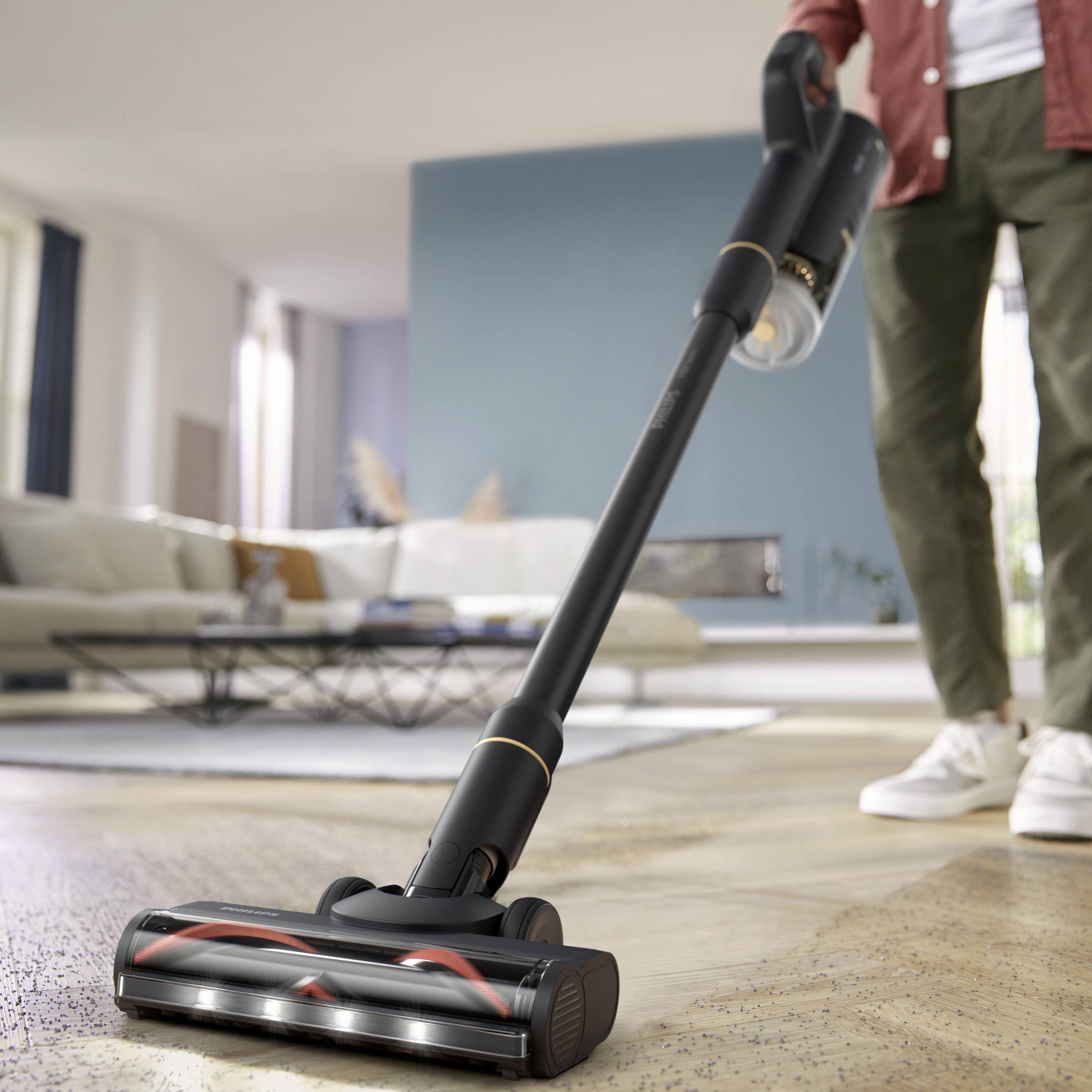 Eine Person benutzt einen kabellosen Staubsauger auf einem Teppich in einem modernen Wohnzimmer mit hellen Fenstern und einem Sofa im Hintergrund.