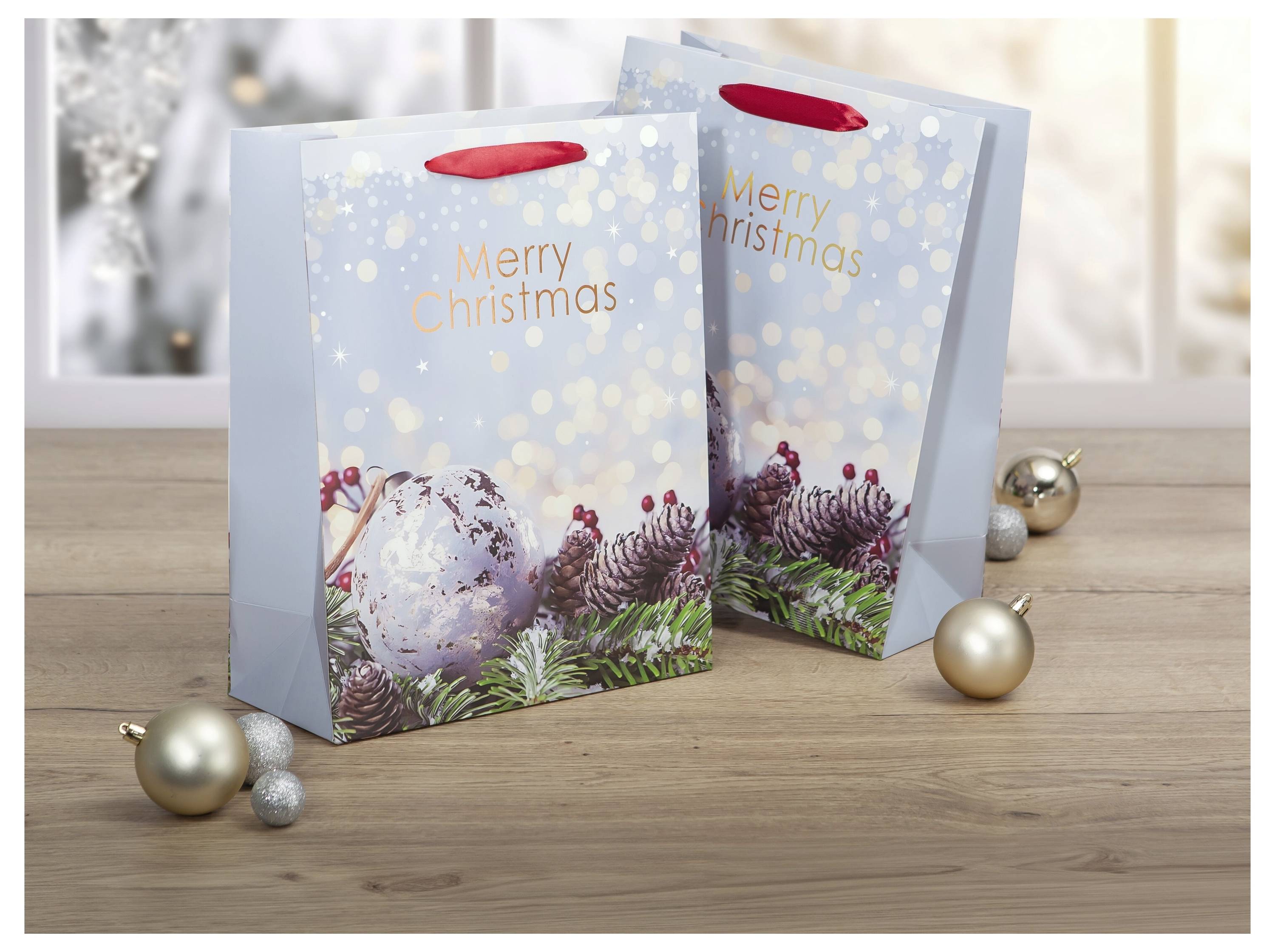 Zwei blaue Weihnachtsgeschenktüten auf einem Holztisch, jede bedruckt mit „Merry Christmas" und einem Winterdesign mit Beeren und Kiefer, mit Weihnachtsornanten in der Nähe.