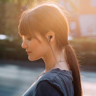 Eine junge Frau mit langen Haaren hört mit kabellosen Ohrhörern Musik. Sie ist im Profil zu sehen, im Hintergrund verschwommene Natur.