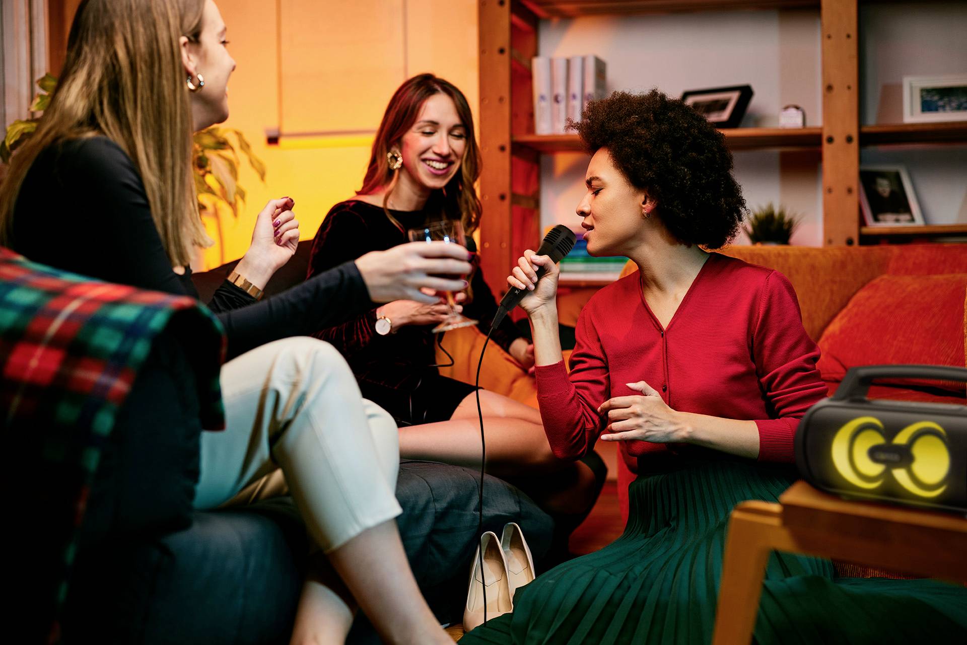 Drei Frauen sitzen in einem Wohnzimmer, trinken Wein und singen Karaoke. Eine Frau hält ein Mikrofon, die Stimmung ist fröhlich und entspannt.