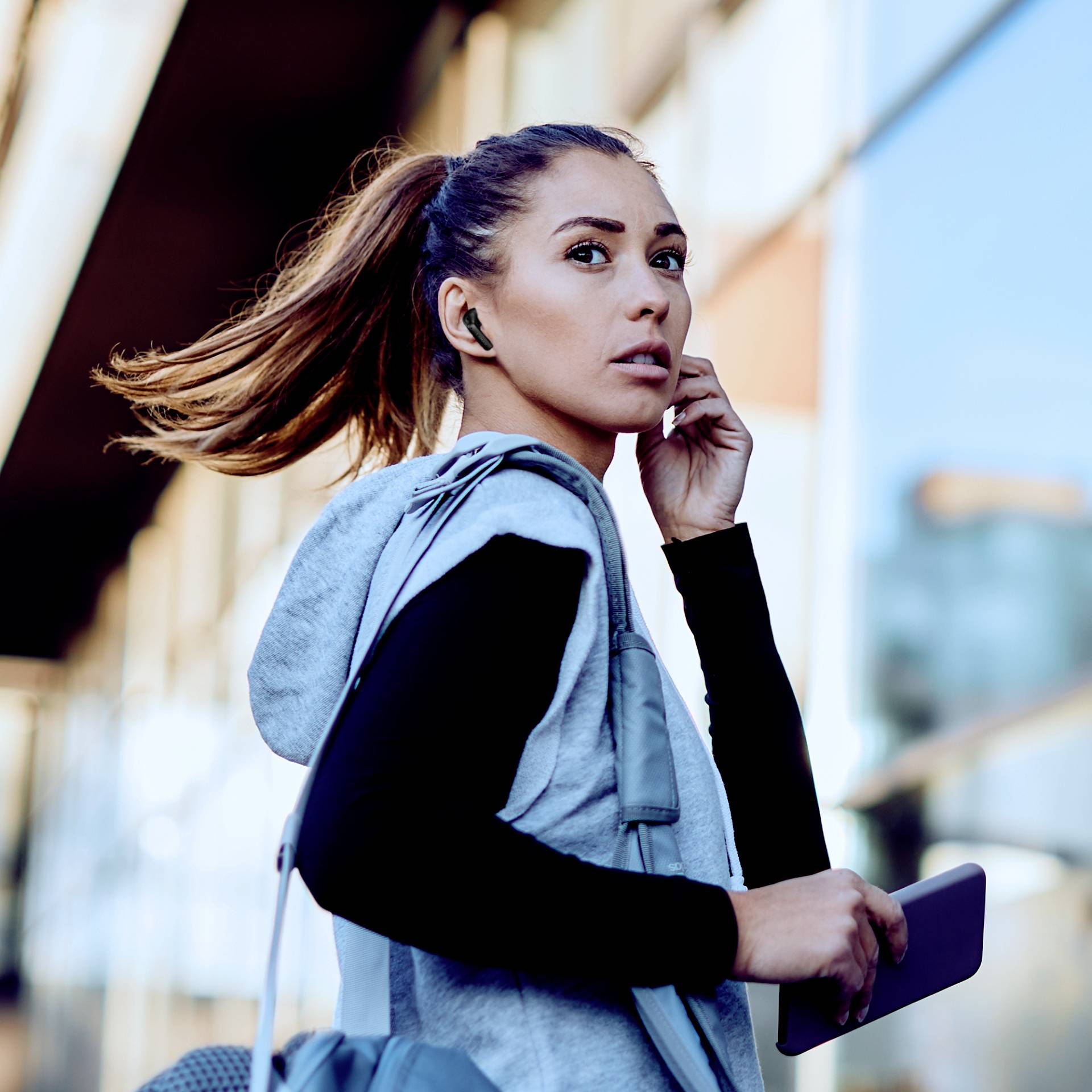 Eine Frau in Sportkleidung hält ein Smartphone, hat Ohrhörer im Ohr und schaut nach links, während sie auf einer Straße geht.