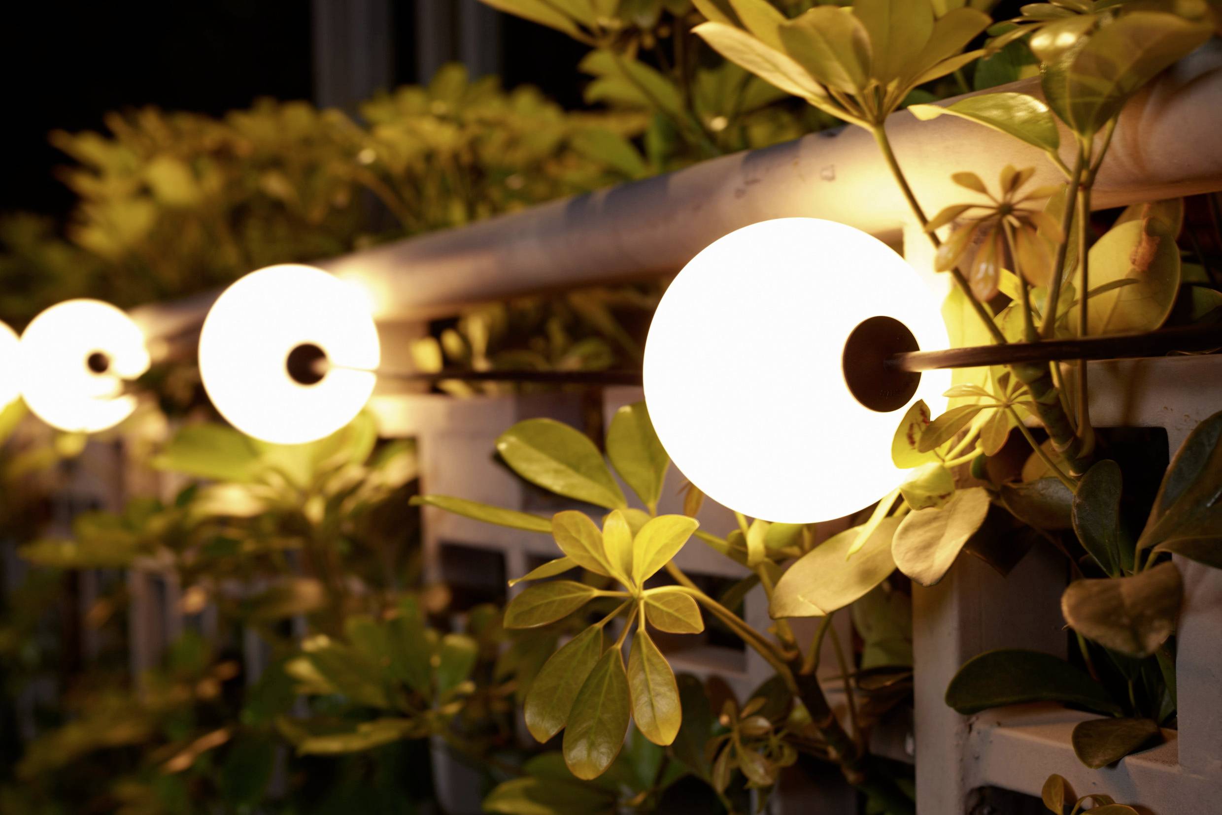 Gartenbeleuchtung mit kreisförmigen Lampen entlang eines Geländers, umgeben von grünem Laub. Warmes und einladendes Ambiente.