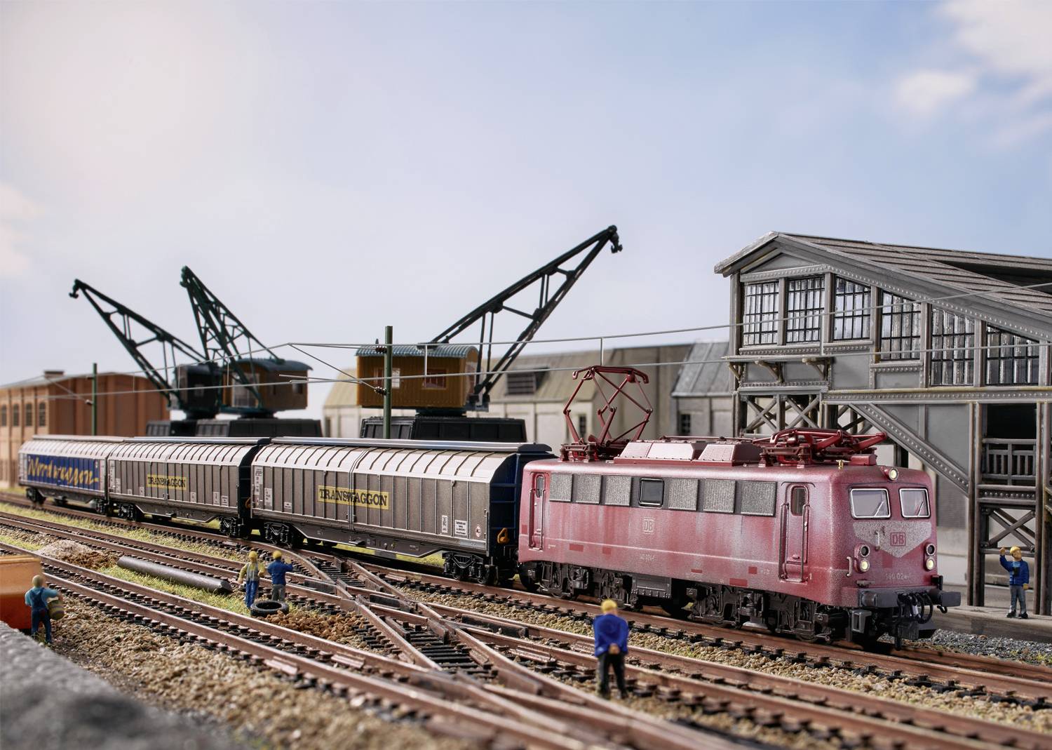 Modelleisenbahn-Szene mit rotem Zug und Waggons auf Bahngleisen. Arbeiter stehen neben Industriegebäuden und Kränen unter blauem Himmel.