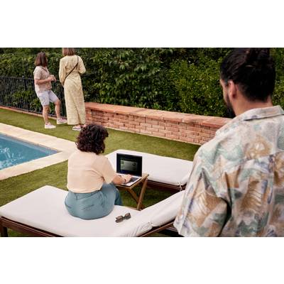 Eine Frau sitzt auf einer Sonnenliege, arbeitet am Laptop. Nah am Pool. Zwei weitere Personen stehen im Hintergrund und schauen weg.