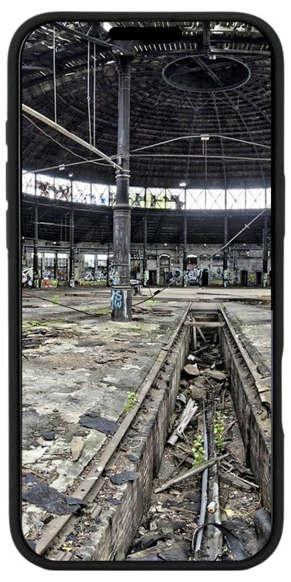 Ein verlassener, verfallener Industriekomplex mit Graffiti an den Wänden. Im Vordergrund sind alte Schienen und Bruchstücke sichtbar.