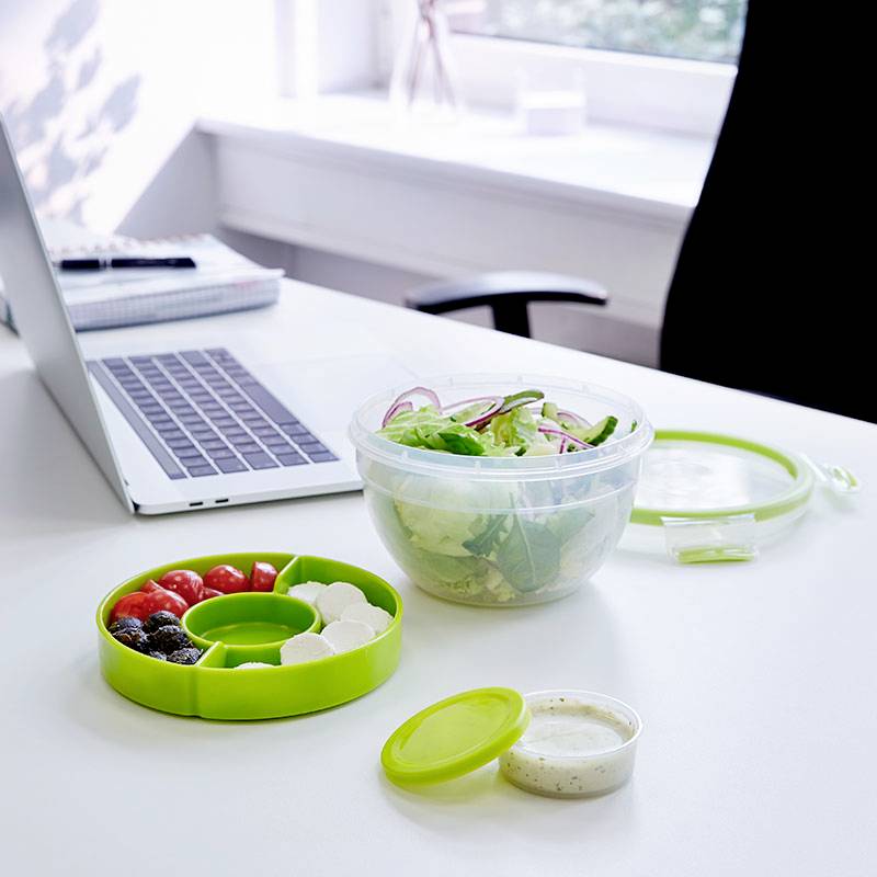 Salatschüssel und Bento-Box auf einem Schreibtisch neben einem Laptop in einem Büro, symbolisieren gesunde Ernährung bei der Arbeit.