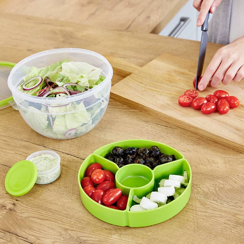 Eine Person schneidet Kirschtomaten auf einem Holzbrett. Daneben stehen eine Schüssel mit Salat und ein Behälter mit geschnittenem Gemüse.