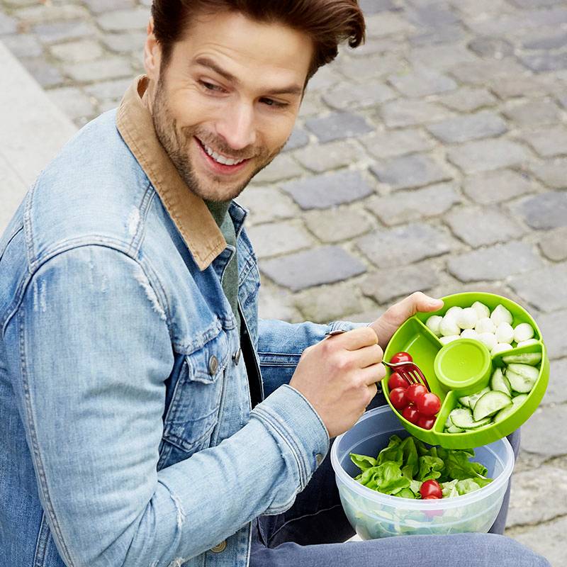 Ein Mann sitzt draußen auf Kopfsteinpflaster und isst aus einem Behälter mit frischem Gemüse wie Tomaten und Gurken.