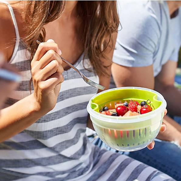 Eine Frau in gestreiftem Kleid hält eine Gabel und isst aus einer Schüssel mit frischem Obstsalat. Im Hintergrund unscharf.