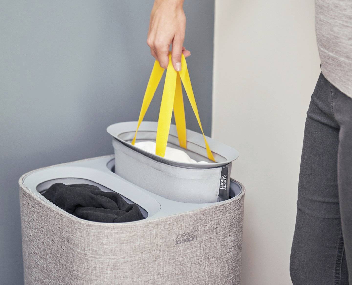 Eine Person hebt einen Einsatz mit gelbem Griff aus einem Doppel-Wäschesammler. Ein schwarzes Kleidungsstück ist im anderen Fach.