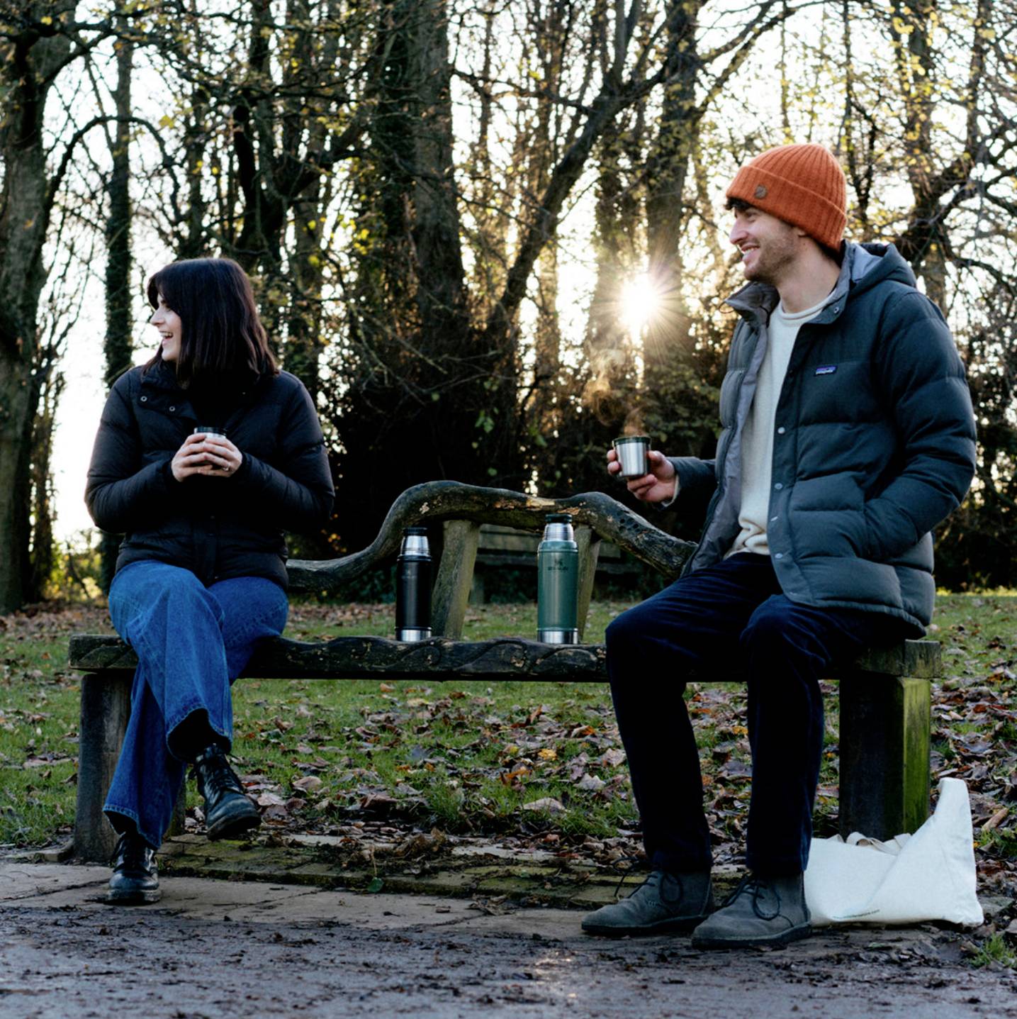 Eine Frau und ein Mann sitzen auf einer Parkbank im Winter. Beide halten Thermosbecher, lächeln und sprechen miteinander.