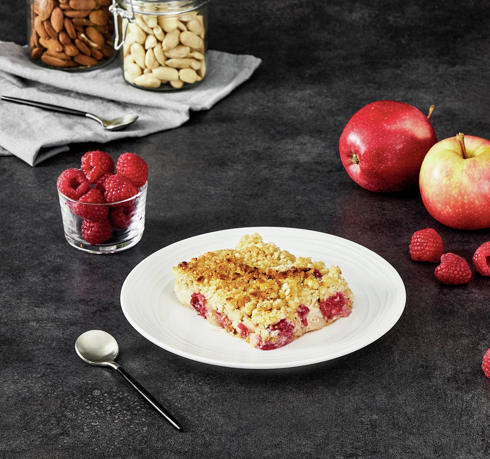 Ein Teller mit einem Stück Himbeer-Streuselkuchen, umgeben von frischen Himbeeren, einem Apfel, einer Gabel und Nüssen im Glas.