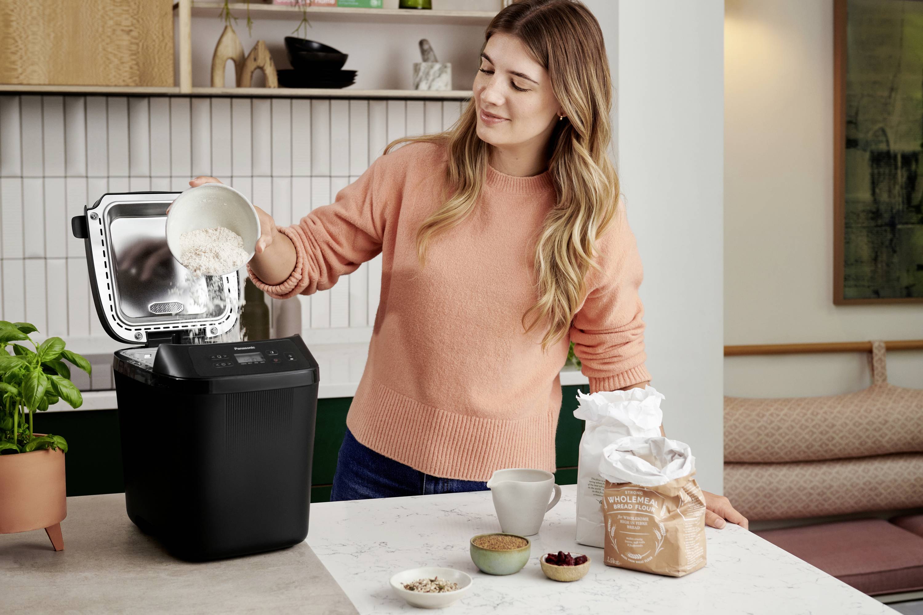 Frau in orangefarbenem Pullover füllt Mehl in einen Brotbackautomaten. Küche mit Pflanzen im Hintergrund.