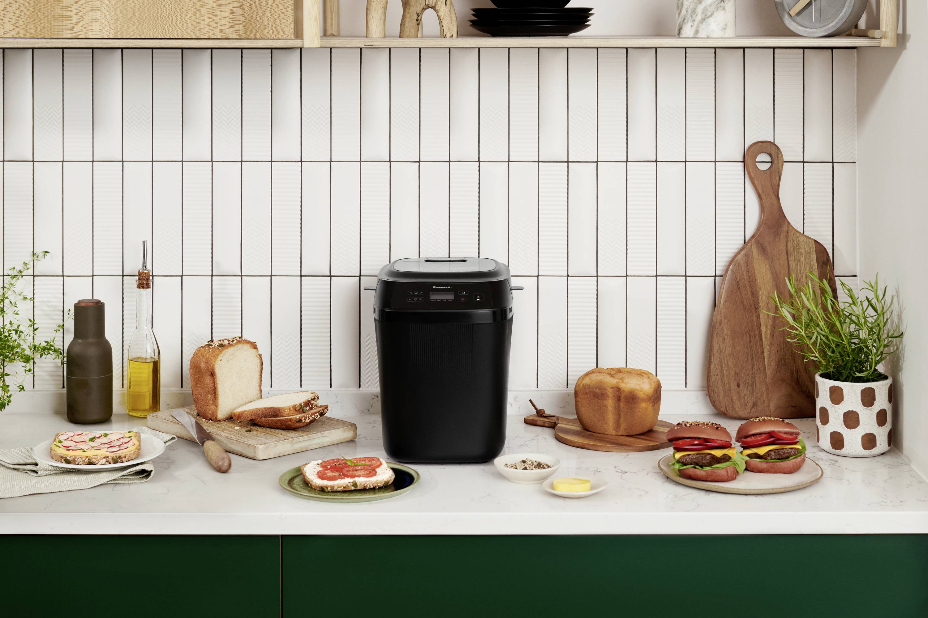 Schwarz Brotbackautomat auf Küchenzeile, umgeben von frischem Brot, Aufschnitt und Pflanzen. Holzbretter und Geschirr dekorieren den Raum.