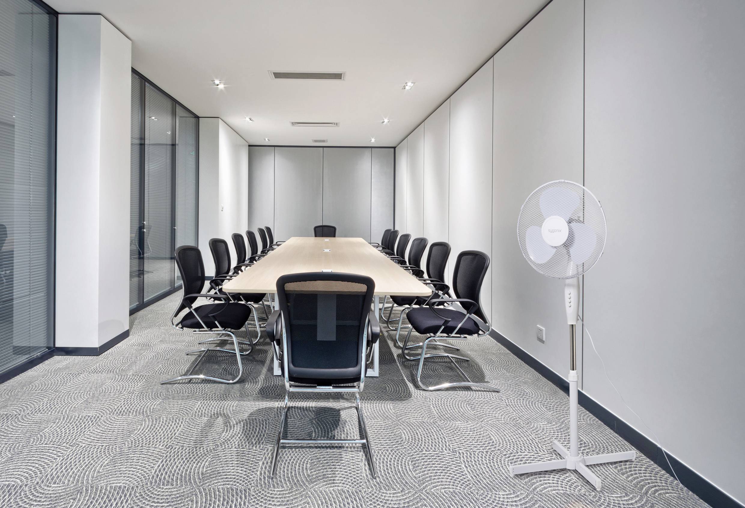 Moderner Konferenzraum mit langem Holztisch, umgeben von schwarzen Bürostühlen. An der Wand steht ein weißer Standventilator.