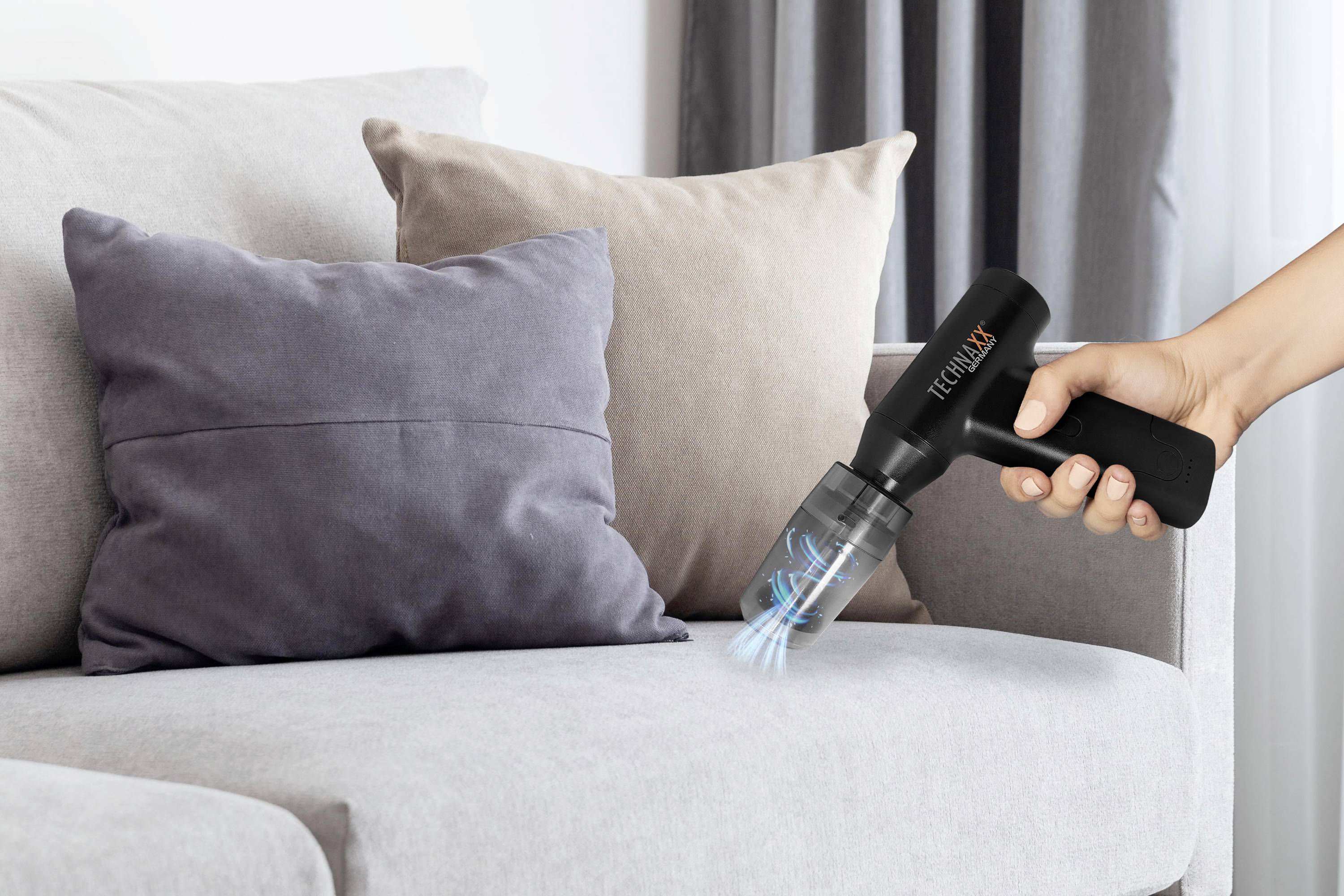 Eine Hand hält einen kleinen, schwarzen Handstaubsauger und reinigt ein graues Sofa. Im Hintergrund sind Kissen sichtbar.
