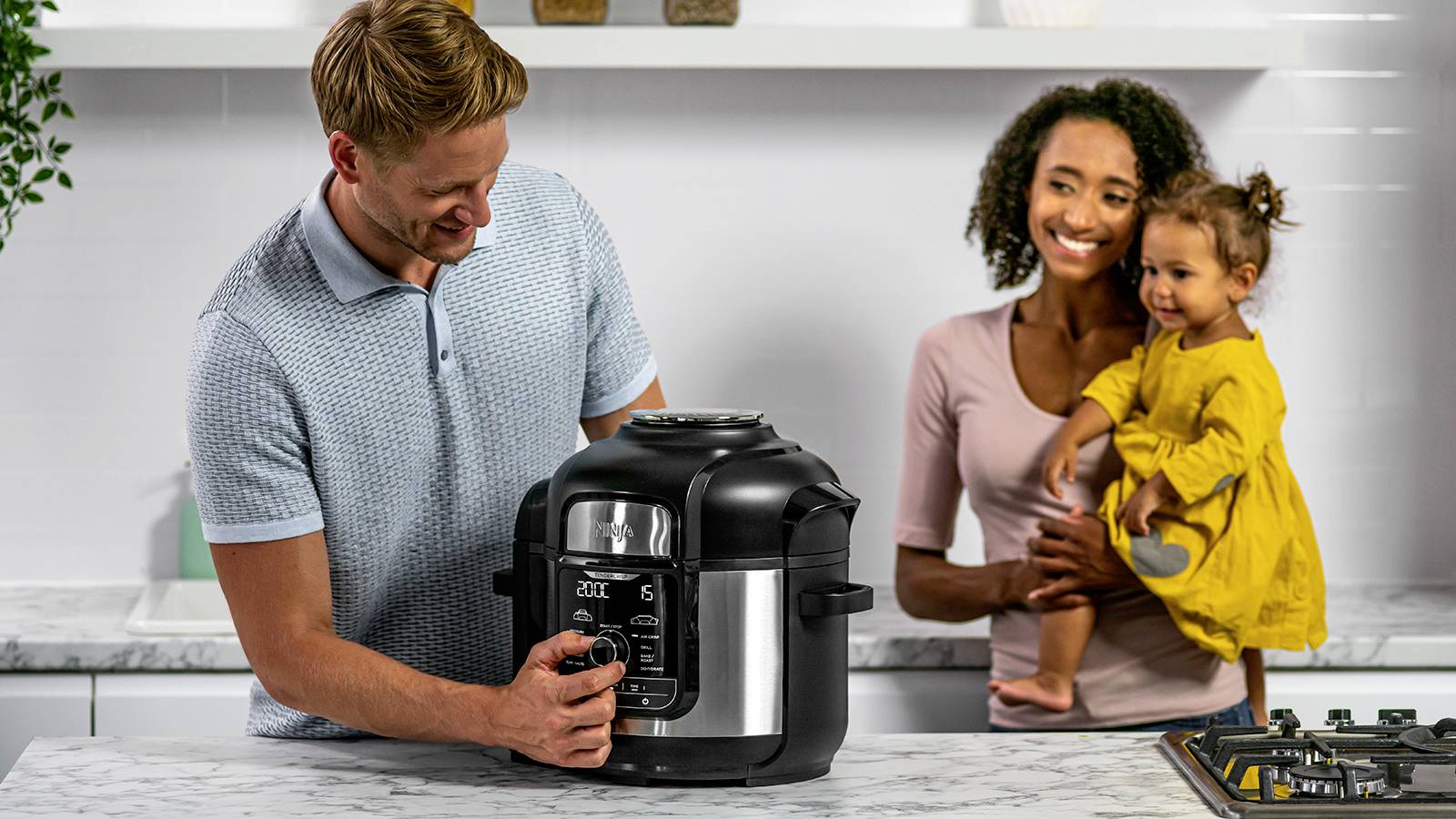 Ein Mann bedient ein Küchengerät auf einer Küchenarbeitsplatte, während eine Frau mit einem Kleinkind im Arm zuschaut und lächelt.