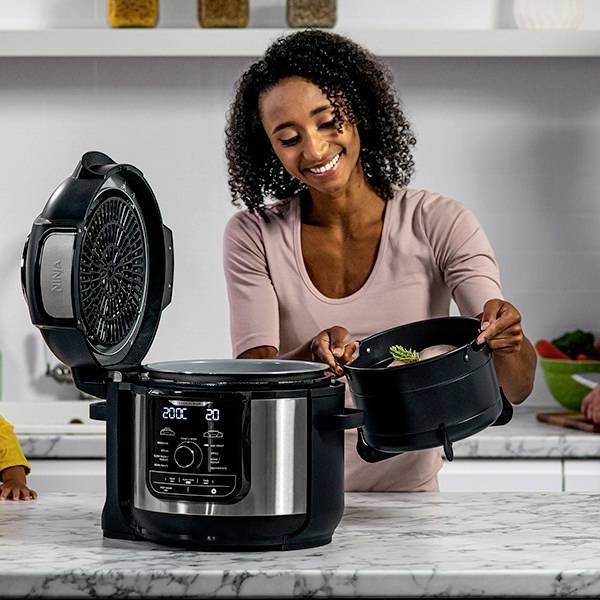 Eine Frau in einer Küche nutzt einen Multifunktionskocher. Sie hält den Deckel. Im Hintergrund stehen Gewürze und Gemüse auf dem Tisch.