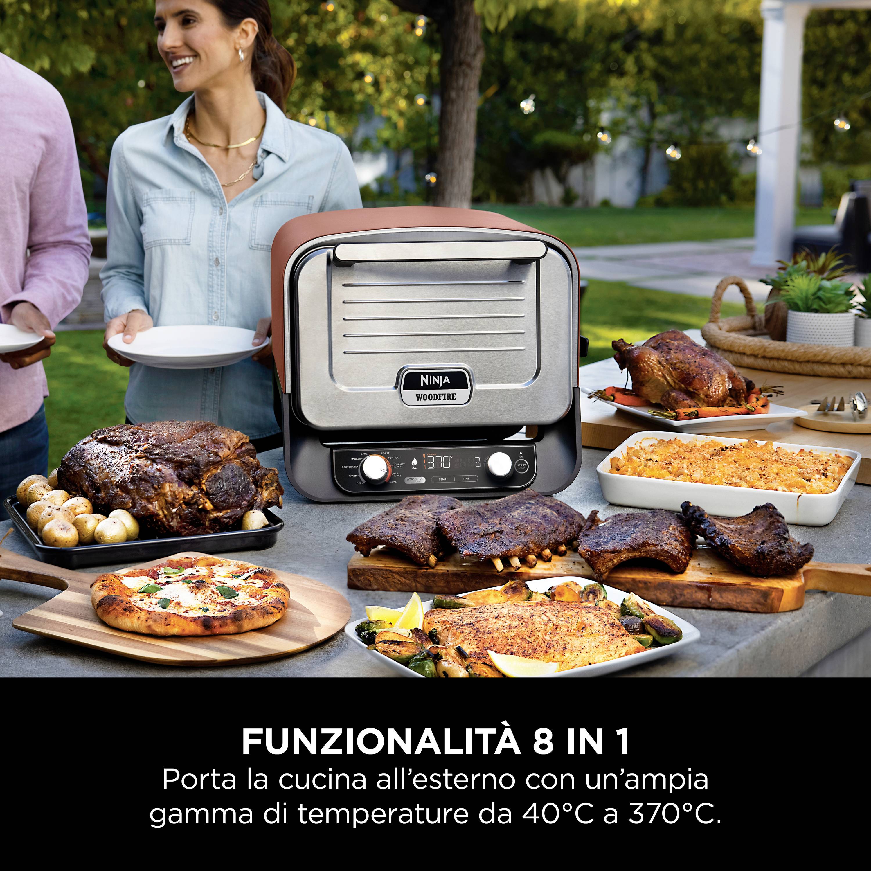 Ein Mann und eine Frau stehen neben einem Tisch im Freien mit einem multifunktionalen Grill. Verschiedene Gerichte sind auf dem Tisch angerichtet.