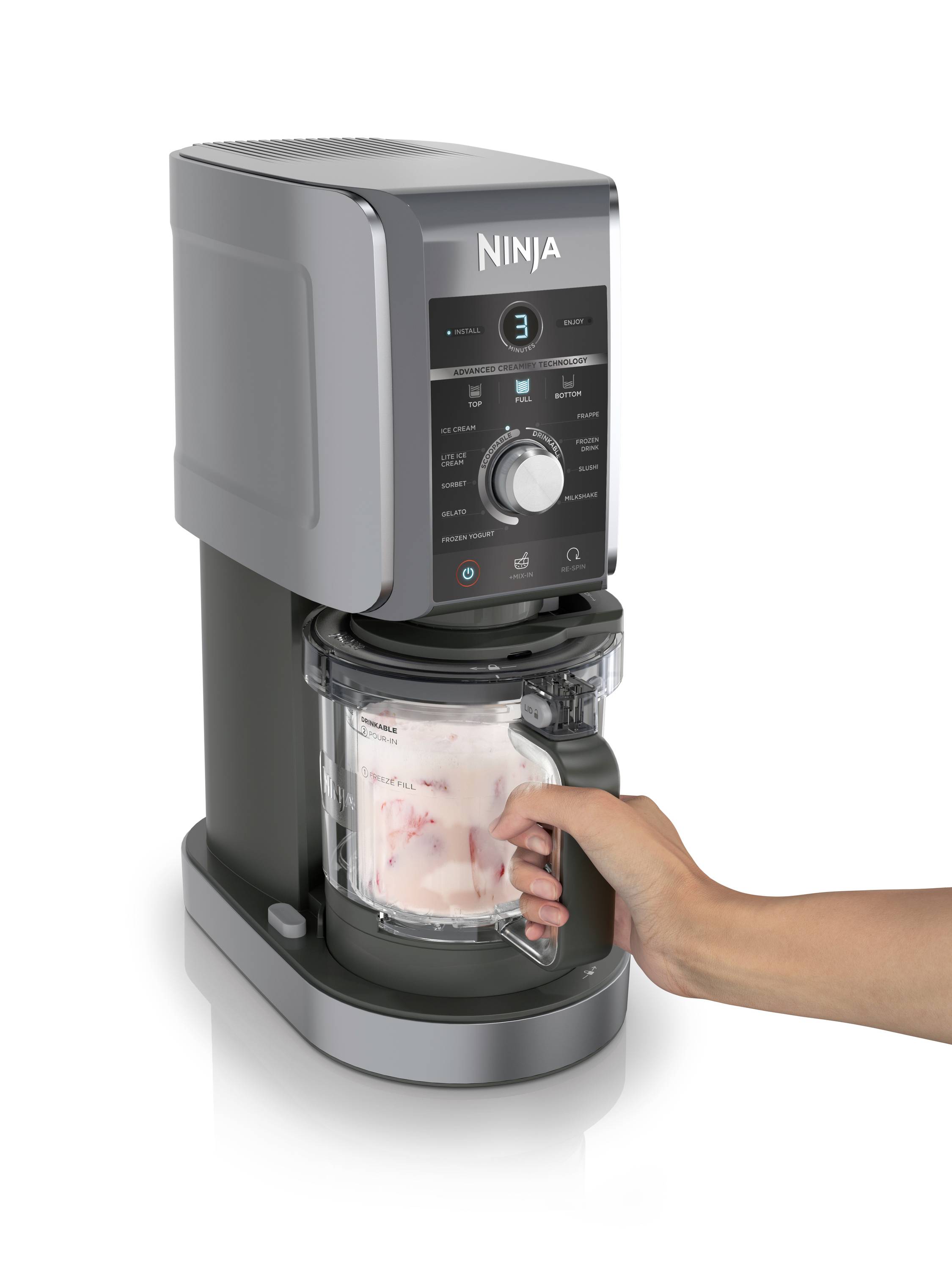 Eine Person hält einen Behälter unter einem grauen Küchengerät, das eine Funktion mit Betriebsanzeige ausführt. Der Behälter enthält gefrorene Zutaten.