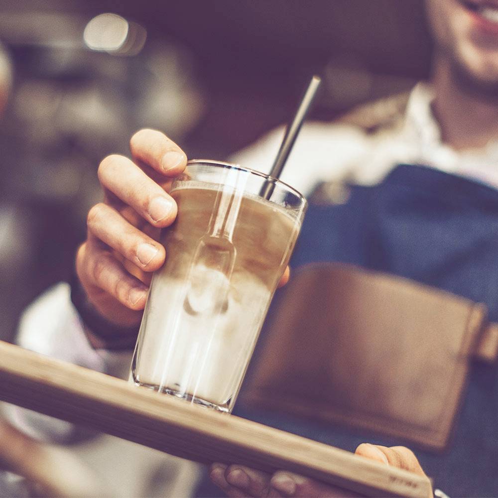 Eine Person hält ein Tablett mit einem Iced Coffee, dekorativem Schaum und Strohhalm. Trägt ein blaues Schürze.