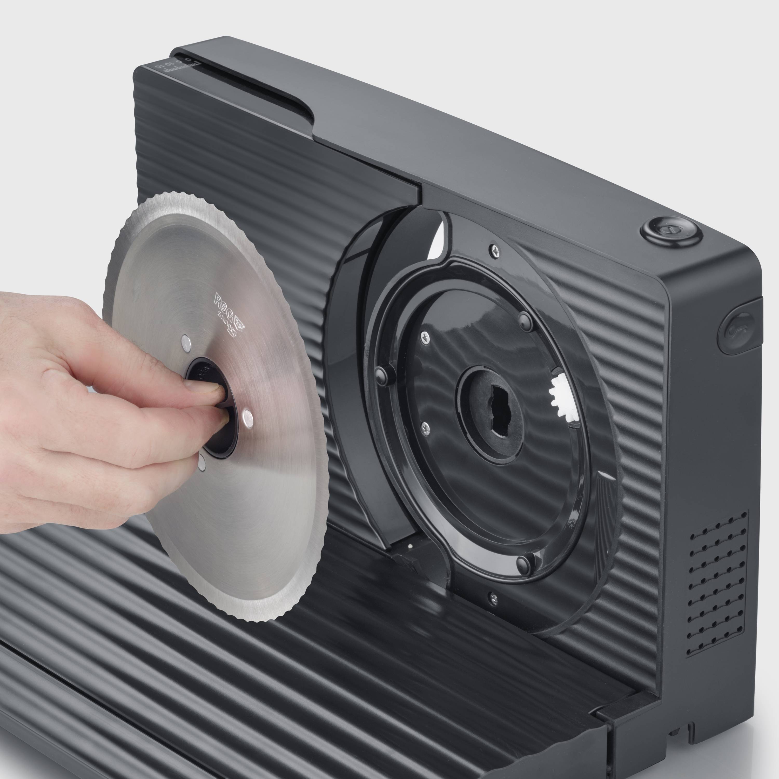 Eine Hand hält eine runde Metallklinge vor einem schwarzen elektrischen Gerät, das wie ein Allesschneider wirkt.