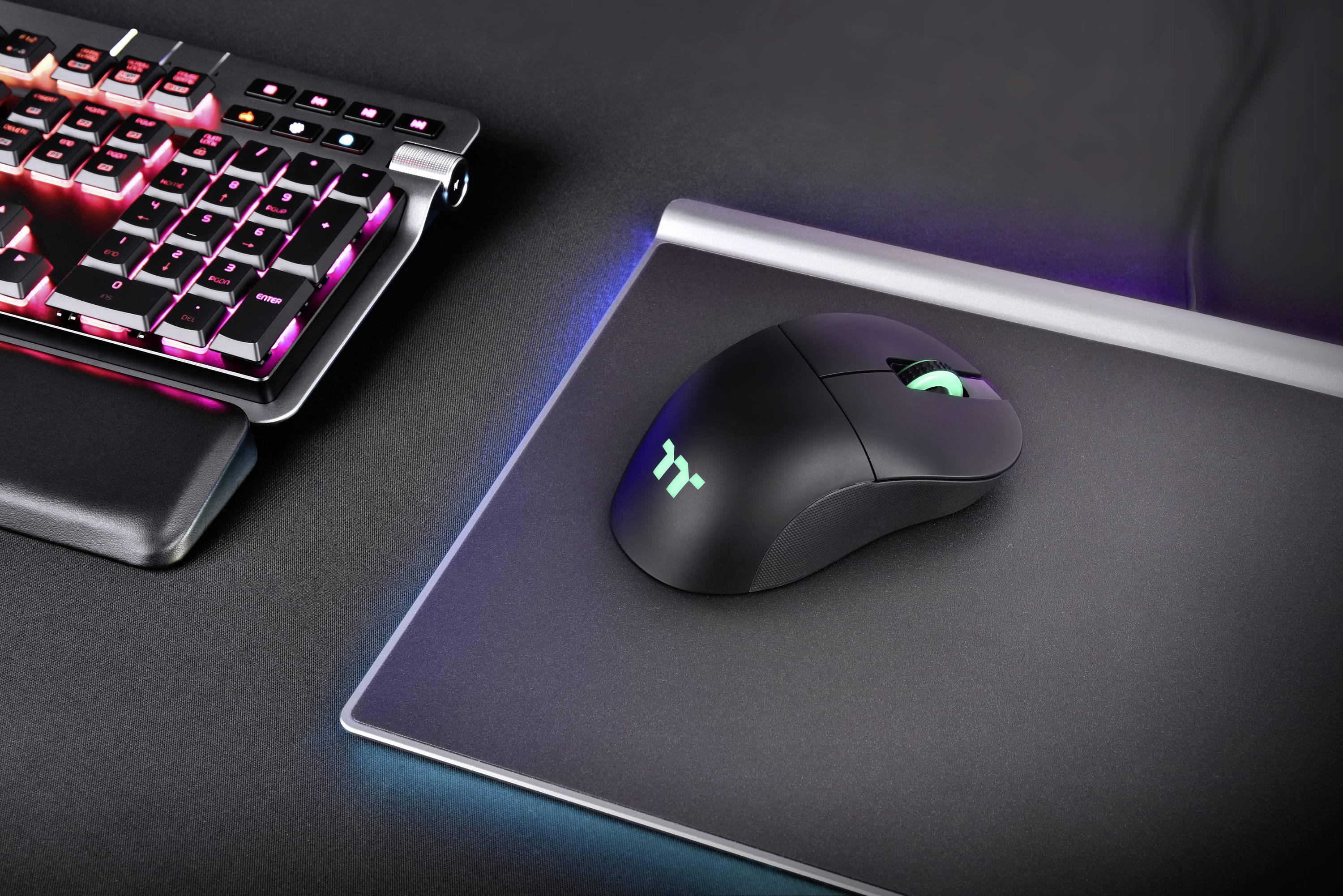 Eine schwarze Gaming-Maus mit leuchtendem Logo auf einem Mousepad neben einer beleuchteten Gaming-Tastatur auf einem Schreibtisch.