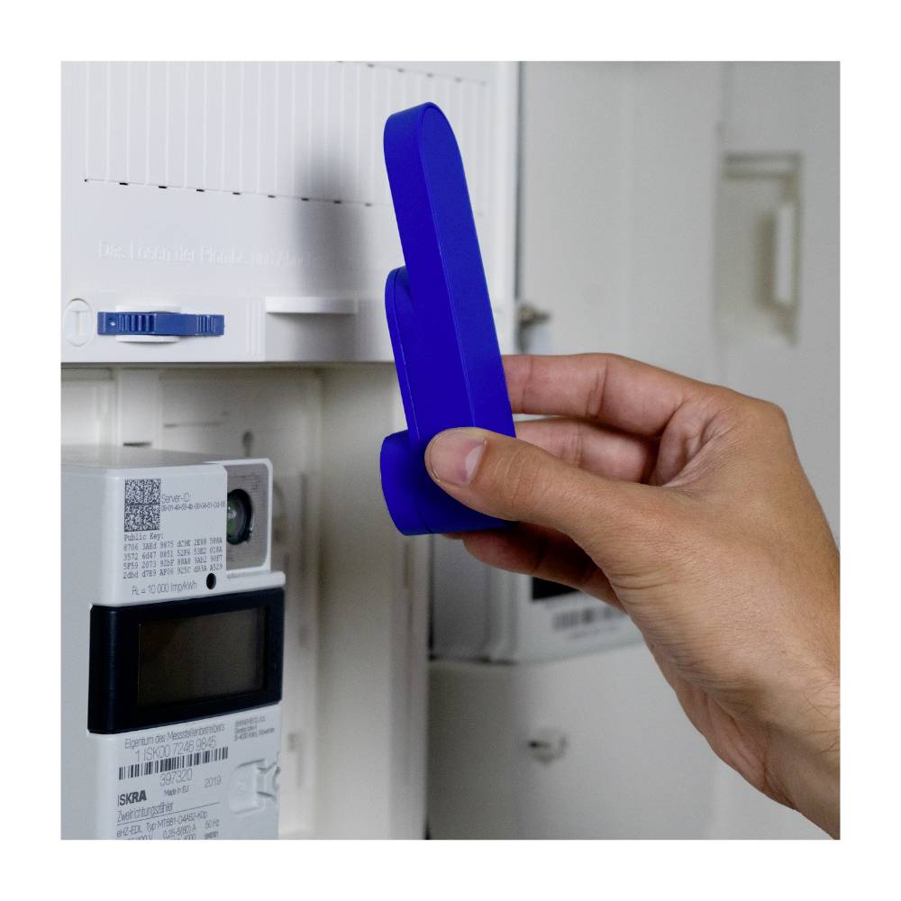 Eine Hand hält ein blaues Gerät vor einen digitalen Stromzähler, erfasst wahrscheinlich Daten.