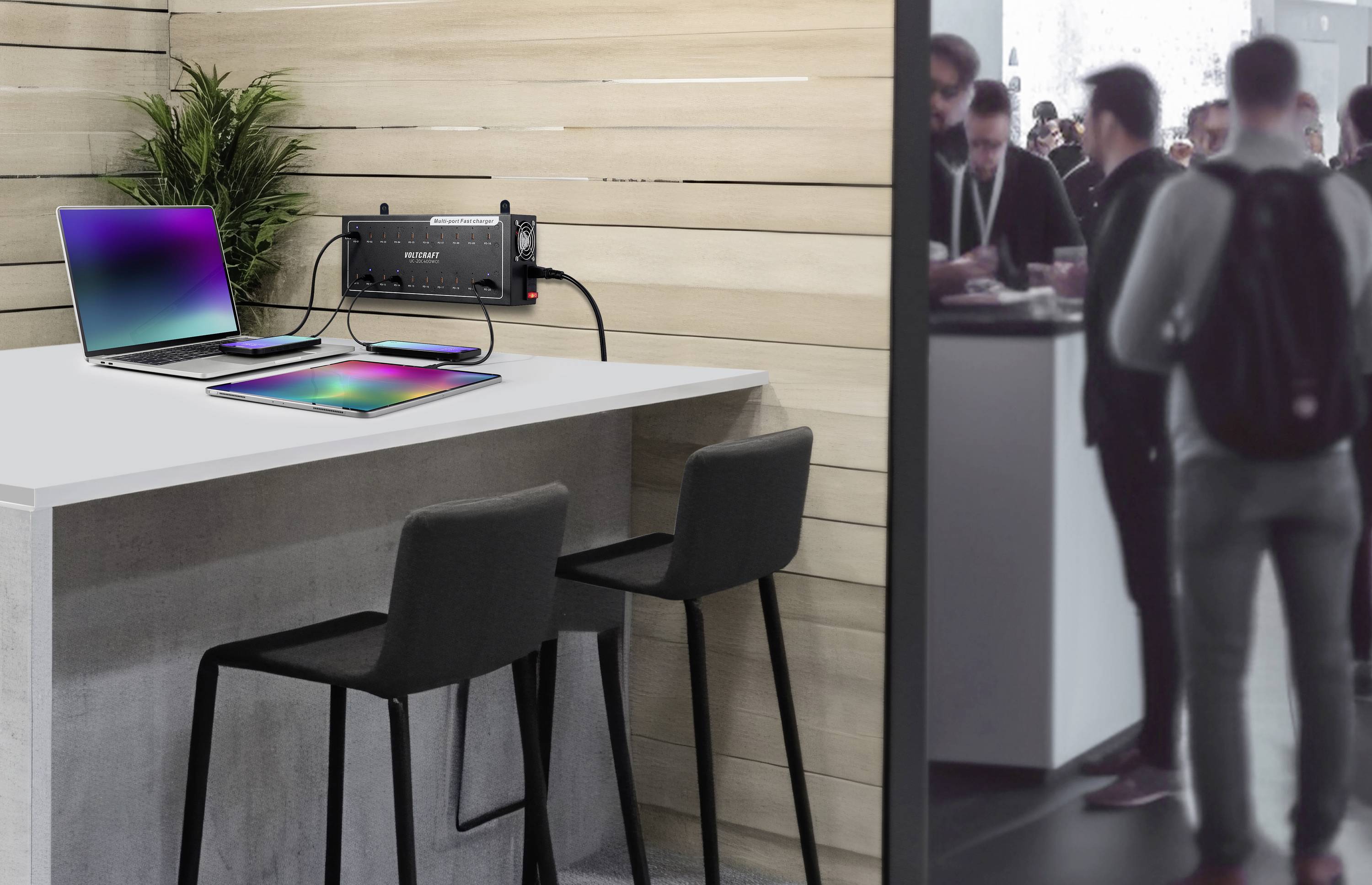 Ein moderner Arbeitsplatz mit zwei Laptops auf einem hohen Tisch, daneben befinden sich barähnliche Stühle. Im Hintergrund Menschen in einem Gespräch.